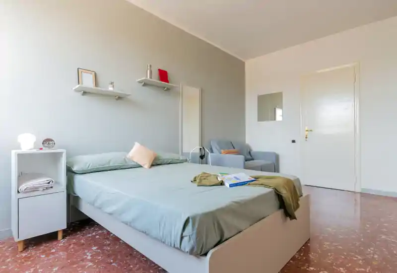 Dormitorio luminoso y bien decorado con cama completa, mesita, estantes y un sillón confortable — ideal para una estancia relajante.