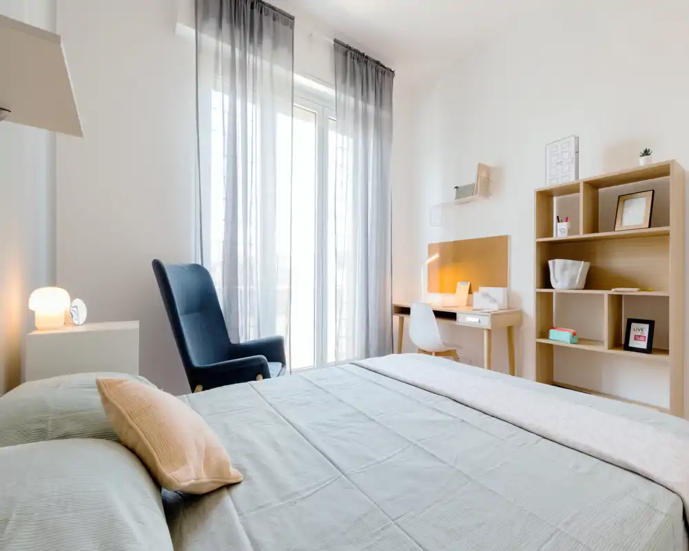 Dormitorio luminoso y moderno con cama bien hecha, un sillón cómodo junto a la gran ventana y un pequeño escritorio con estantería — ideal para relajarse o trabajar ligeramente.