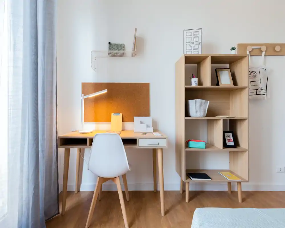 Rincón de dormitorio luminoso y minimalista con escritorio sencillo, tablero de corcho y estantería; luz natural suave y lámpara de escritorio que crean un espacio de trabajo tranquilo junto a la cama.