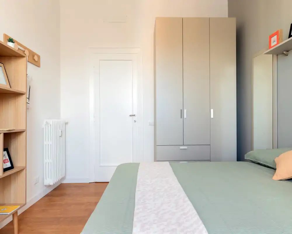 Dormitorio luminoso y moderno con cama bien hecha, armario neutro y estantería de madera — ambiente tranquilo y acogedor.