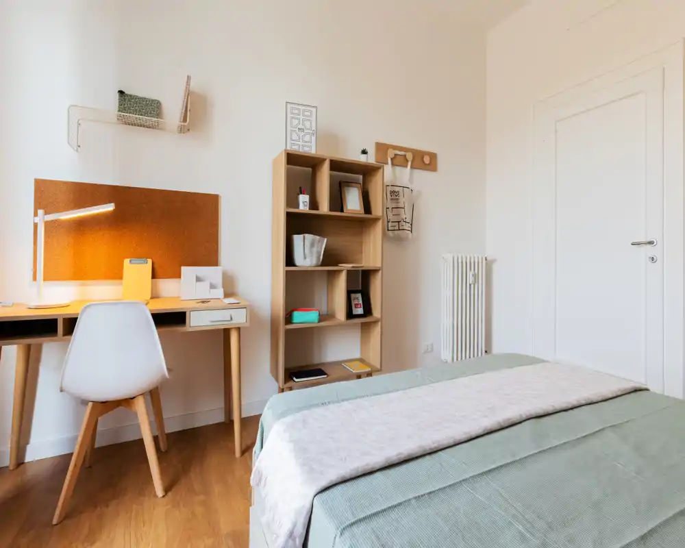 Dormitorio luminoso y bien decorado con la cama en primer plano, una estantería de madera y un pequeño escritorio con luz cálida para trabajar — ideal para una estancia cómoda o teletrabajo.