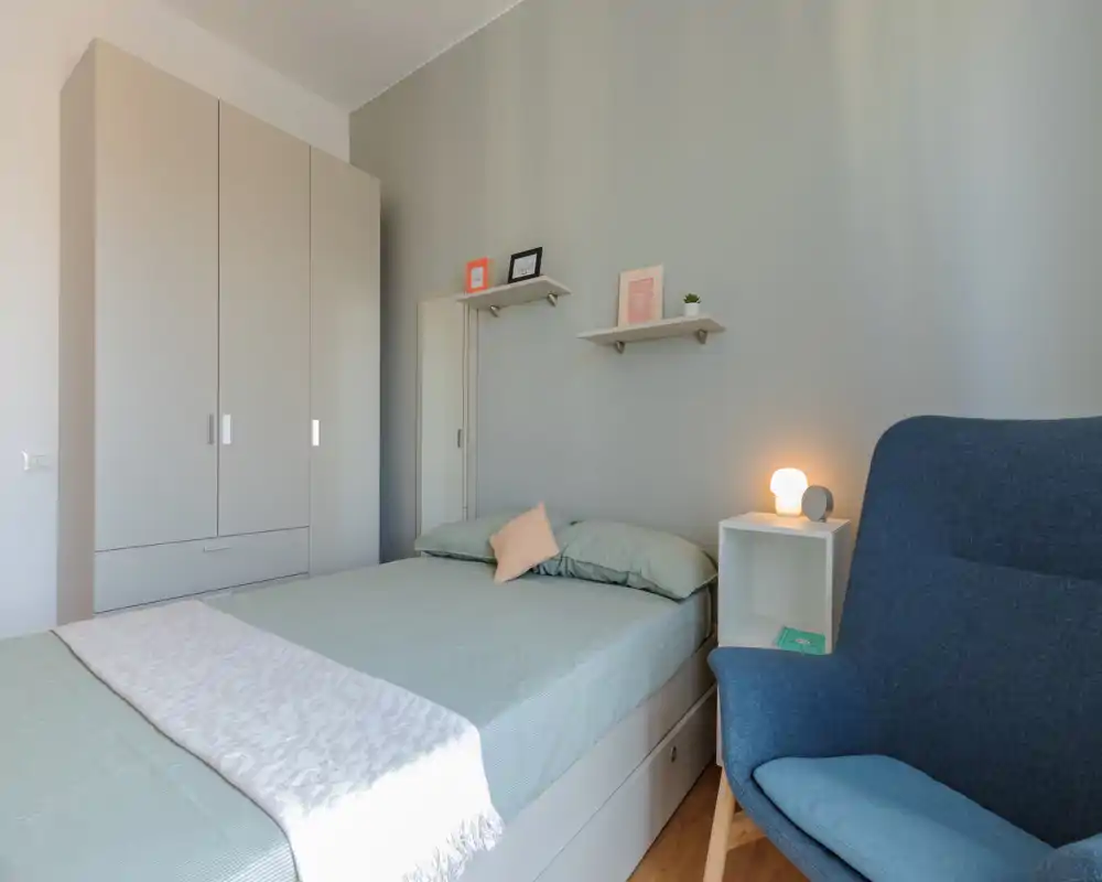 Dormitorio luminoso y bien arreglado con cama doble, ropa de cama verde suave, una estantería de mesilla y un cómodo sillón azul — perfecto para relajarse.
