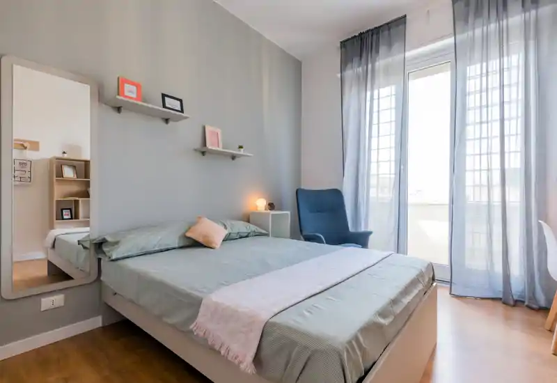 Dormitorio moderno y luminoso con cama doble bien hecha, textiles suaves, un sillón acogedor y mucha luz natural procedente de la ventana balcón.