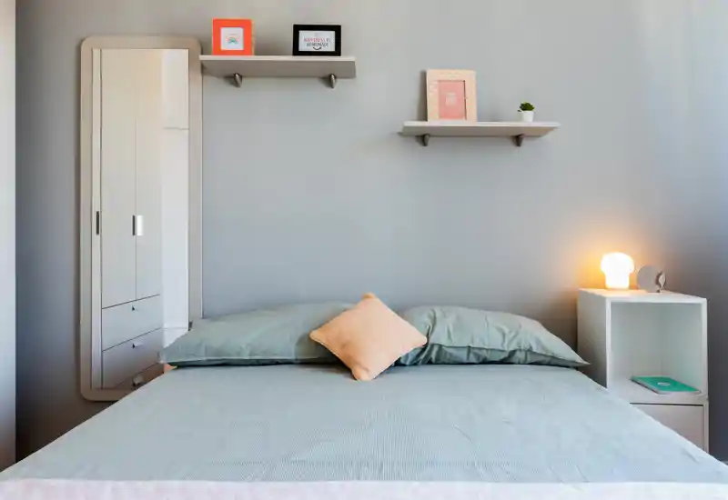 Dormitorio bien decorado con cama doble ordenada, ropa de cama suave, cojín decorativo y una lámpara de mesita que crea un ambiente cálido y moderno.