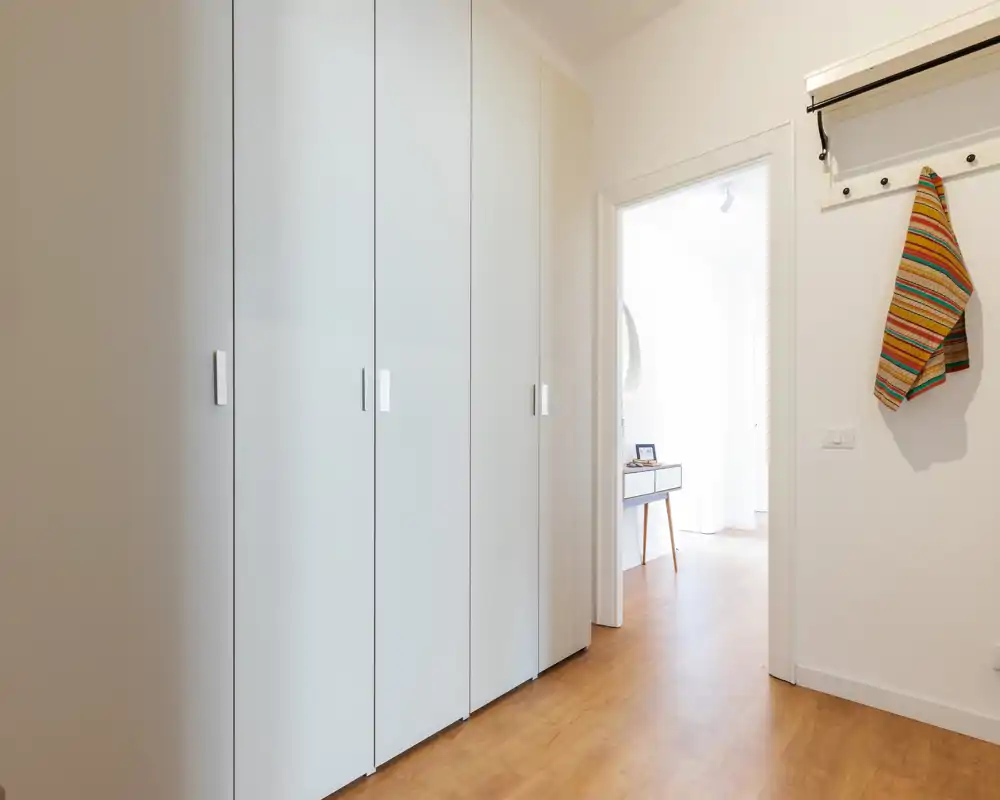 Entrada luminosa y moderna con armarios empotrados blancos, suelo de madera y perchero; estilo limpio y minimalista con luz natural desde la habitación contigua.