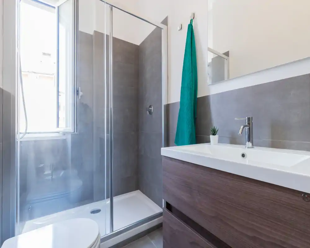 Baño moderno con mampara de ducha de vidrio, mueble con lavabo y luz natural procedente de una ventana: limpio, luminoso y funcional.