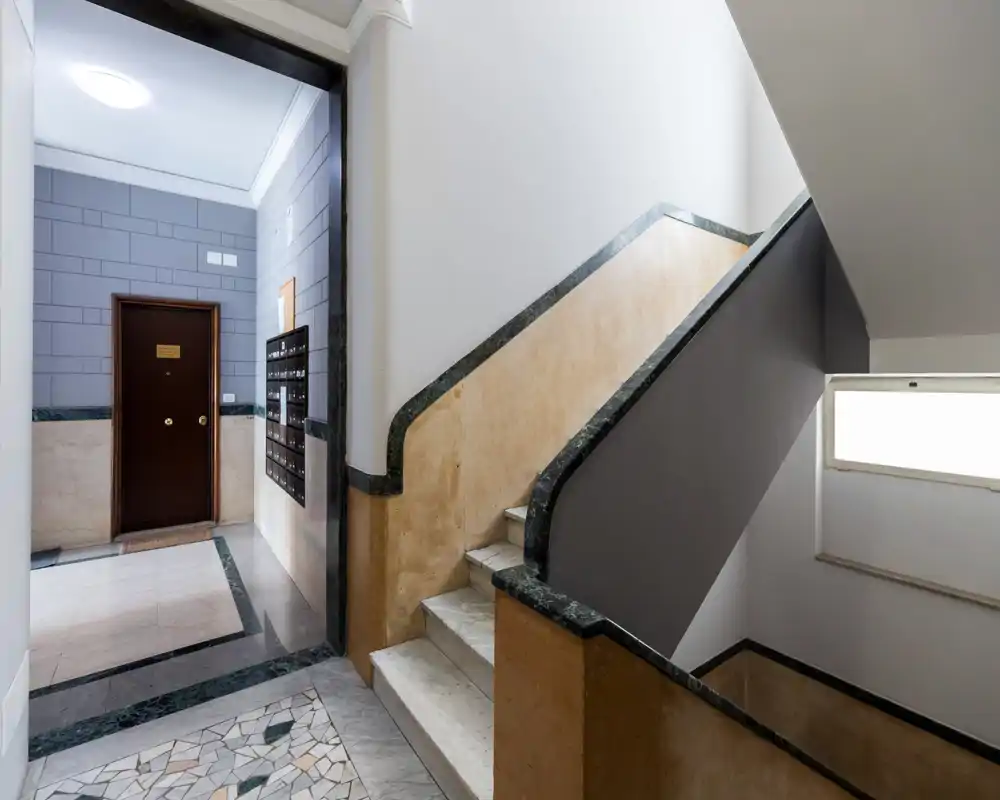 Entrada del edificio y escalera bien iluminadas con peldaños de mármol, suelo decorativo y buzones — zona común limpia y segura.