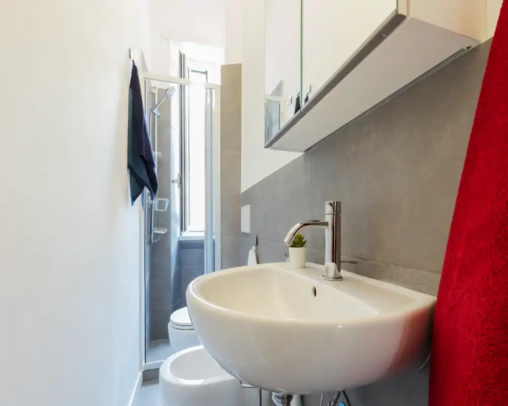 Baño moderno y compacto con lavabo suspendido, grifo cromado y ducha visible al fondo; azulejos grises neutras y luz natural brillante crean una sensación limpia y contemporánea.