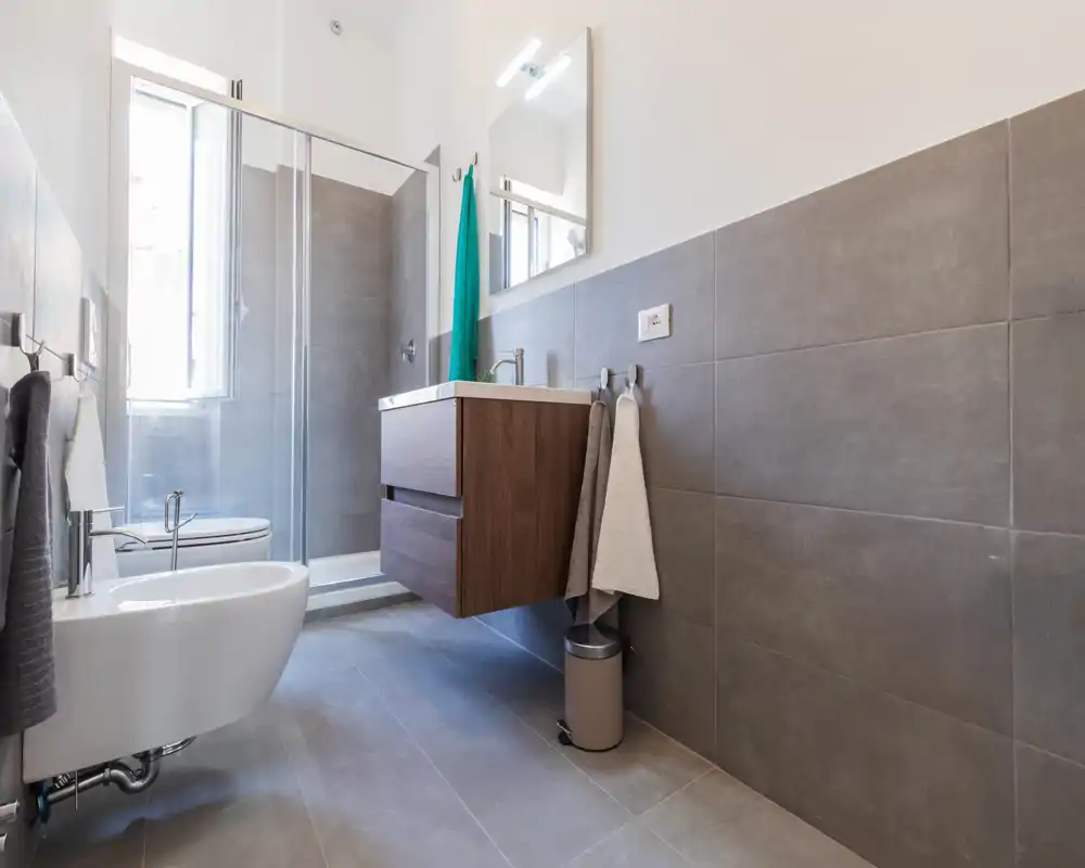 Baño moderno con mampara de ducha, mueble lavabo suspendido y bidé; azulejos grises y abundante luz natural.