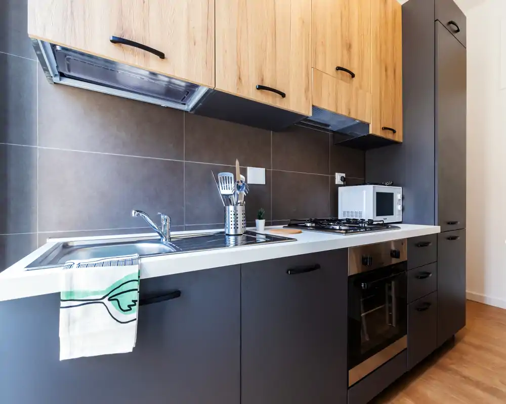 Cocina compacta moderna con muebles bajos oscuros y armarios altos de madera, horno empotrado, placa de gas y fregadero de acero inoxidable — luminosa, bien decorada y lista para usar.