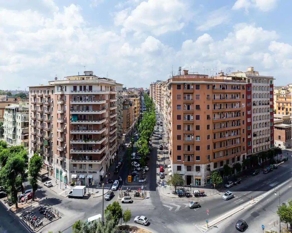 Amplia vista urbana tomada desde un punto elevado, con dos bloques residenciales, una avenida arbolada y una intersección concurrida bajo un cielo parcialmente nublado.
