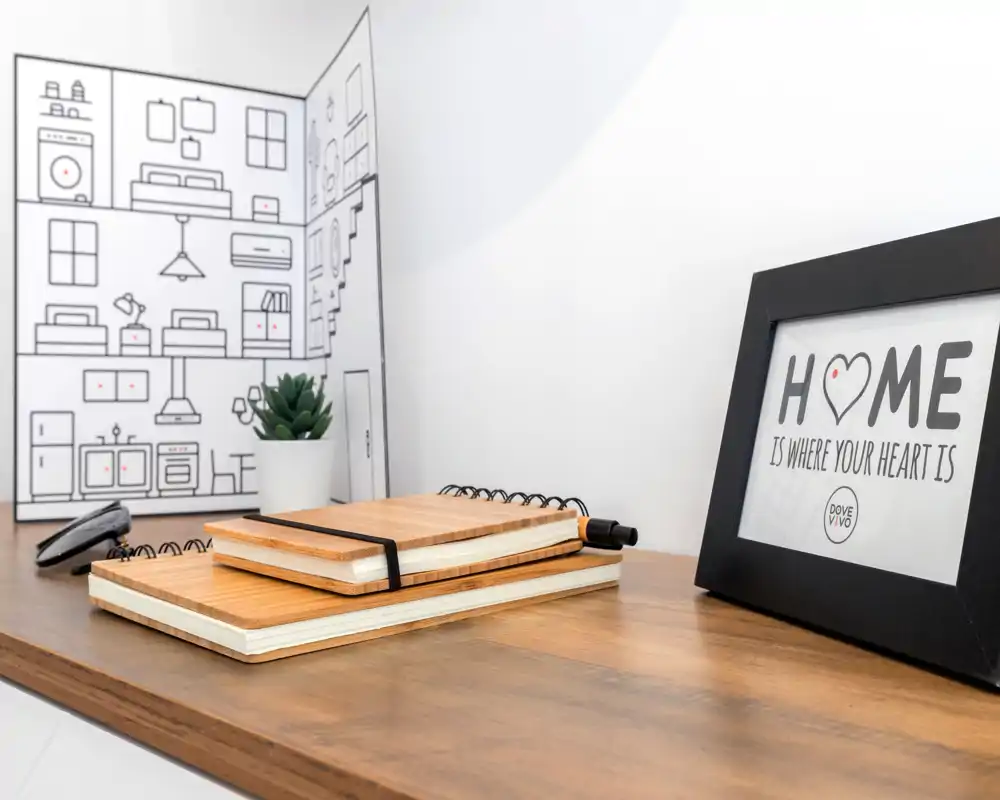 Plano cercano de un rincón de escritorio en la zona de estar: superficie de madera con cuadernos apilados, un marco con la palabra "HOME" y una pequeña planta — estilo limpio y moderno, ideal para anuncios.