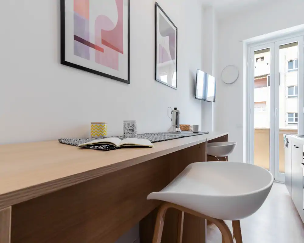 Kitchenette moderna y luminosa con encimera de madera, taburetes y puerta al balcón que aporta luz natural — ideal para vida en ciudad en espacios reducidos.