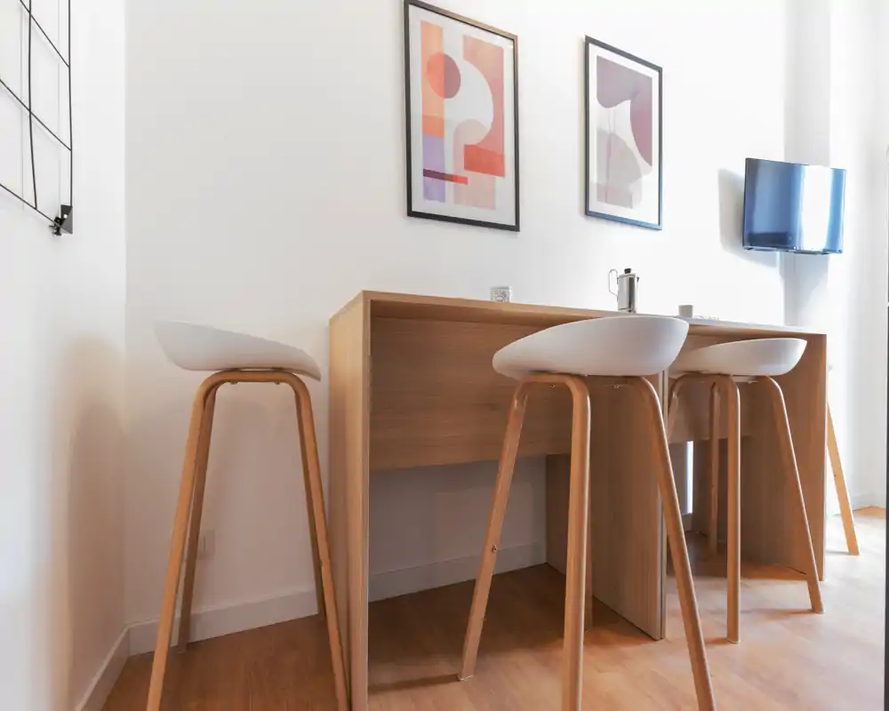 Rincón de cocina moderno y luminoso con una barra de madera y tres taburetes elegantes; decoración minimalista y suelos de madera cálidos que crean un espacio acogedor y funcional.