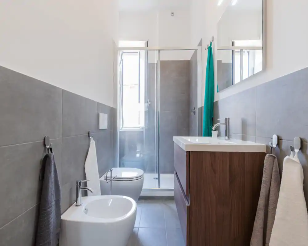 Baño moderno y bien iluminado con ducha, inodoro y bidé suspendidos y mueble lavabo de madera con espejo. Baldosas grises neutras y líneas limpias para un aspecto contemporáneo ideal para anuncios.