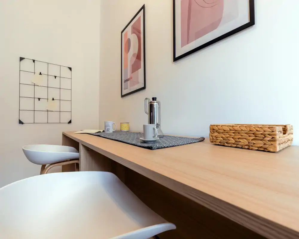 Zona de desayuno minimalista con dos taburetes modernos, encimera de madera, set de café y láminas decorativas — un rincón compacto y luminoso para comidas informales o trabajo.