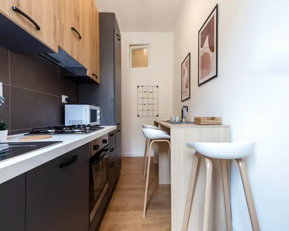 Cocina compacta y moderna con horno empotrado y hornilla de gas, muebles en madera clara y una barra estrecha con taburetes — espacio luminoso y bien decorado, ideal para estancias individuales o en pareja.