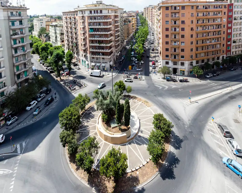 Vista aérea de una rotonda urbana con árboles y edificios de apartamentos alrededor a plena luz del día, ideal para mostrar el contexto del barrio y el trazado de las calles.