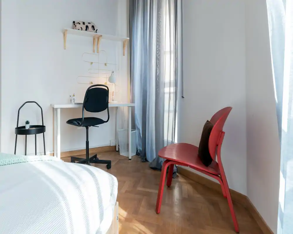 Rincón de dormitorio luminoso y moderno con el borde de la cama visible, un escritorio y una silla roja como punto focal: ambiente limpio y acogedor.