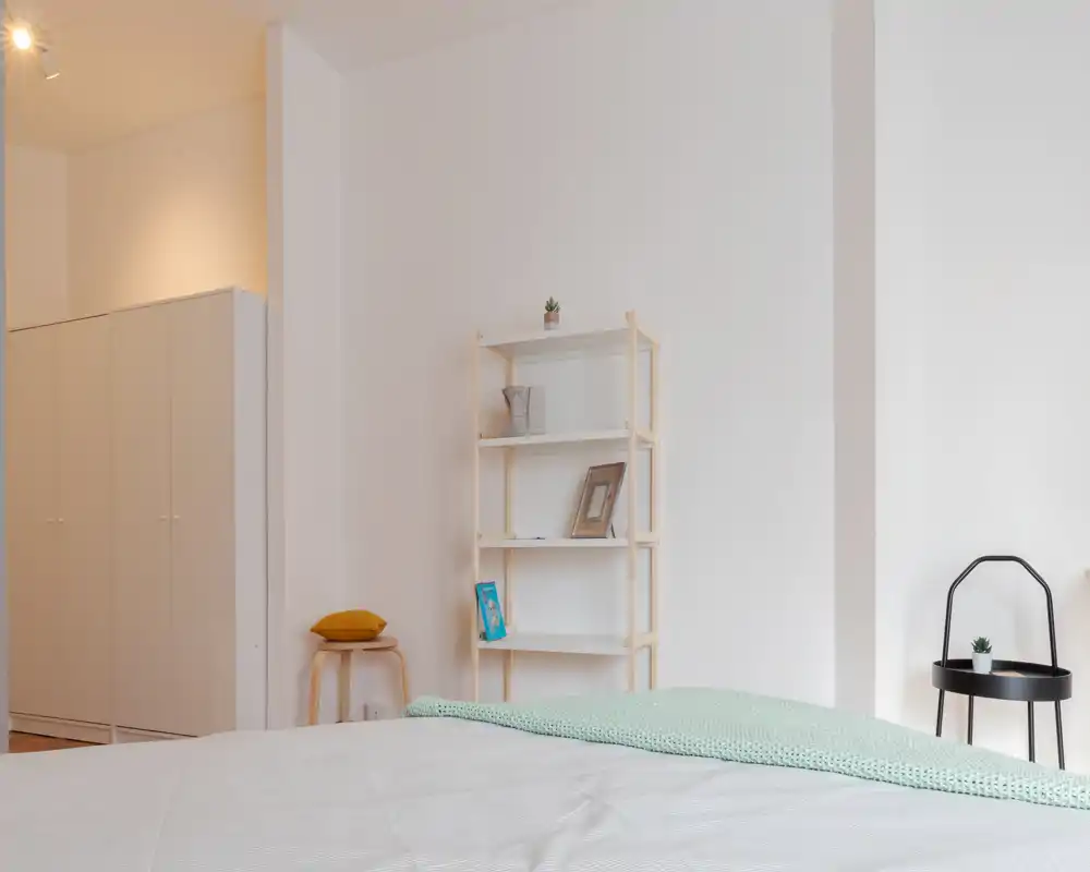 Dormitorio minimalista y luminoso con cama bien hecha en primer plano, manta verde claro y estantería sencilla contra una pared blanca—espacio para dormir claro y tranquilo.