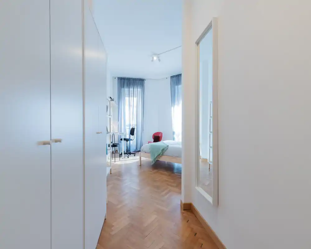 Dormitorio luminoso y aireado con suelo de parquet, cama bien hecha con manta en tonos pastel, escritorio y silla junto a dos grandes ventanas que aportan mucha luz natural.