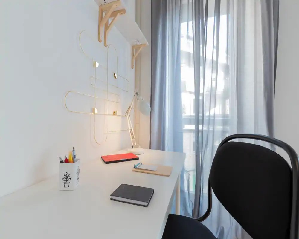 Rincón de oficina en casa luminoso con escritorio blanco, lámpara de sobremesa, cuadernos y una silla cómoda junto a una ventana con cortina traslúcida — ordenado, minimalista y bien iluminado para trabajar o estudiar.