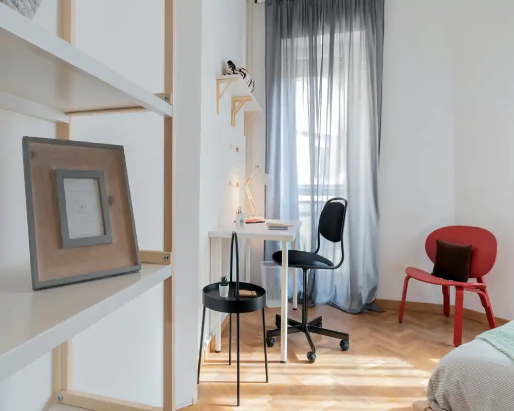 Rincón de dormitorio luminoso y minimalista con un escritorio junto a la ventana, sillas modernas y una pequeña muestra de la cama; suelos de parquet cálidos y textiles acogedores.
