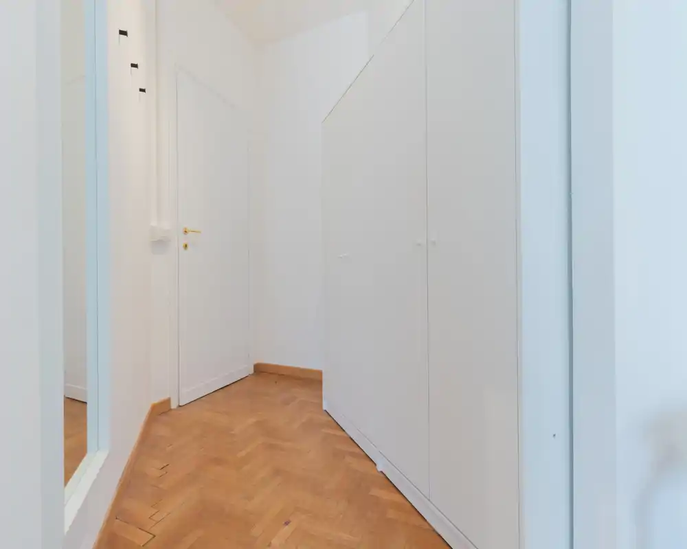 Entrada luminosa y minimalista con armarios empotrados blancos, puerta blanca y suelo de parquet cálido — un área de acceso limpia y funcional.
