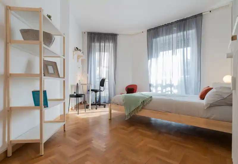 Dormitorio luminoso y minimalista con cama de madera, manta acogedora y grandes ventanas que dejan entrar abundante luz natural.