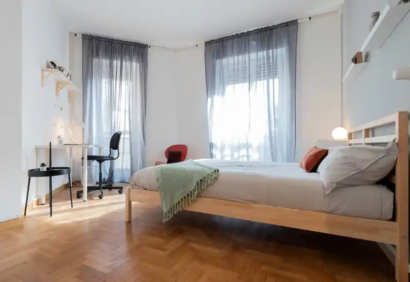 Dormitorio luminoso y minimalista con cama doble de madera, ropa de cama suave y grandes ventanas que dejan entrar luz natural. Incluye un pequeño escritorio y detalles acogedores.