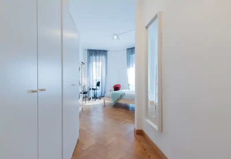 Dormitorio luminoso y aireado con suelo de parquet, cama bien hecha con manta en tonos pastel, escritorio y silla junto a dos grandes ventanas que aportan mucha luz natural.