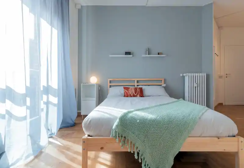 Dormitorio luminoso y aireado con una cama de madera sencilla, ropa de cama suave y una manta mint. Gran ventana con cortinas translúcidas que dejan entrar abundante luz natural para una atmósfera tranquila y acogedora.