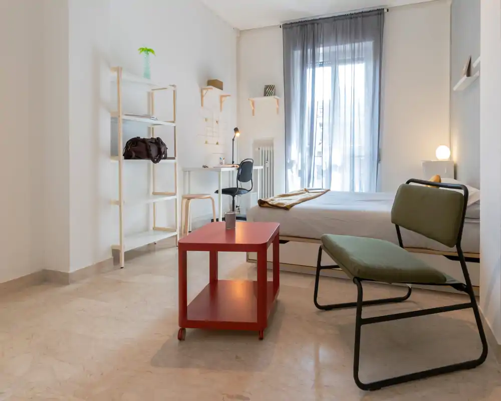 Camera da letto luminosa e minimalista con un comodo letto matrimoniale, una piccola scrivania vicino alla finestra e sedute moderne. Toni neutri e luce naturale creano un’atmosfera calma e ariosa, ideale per affitti brevi.