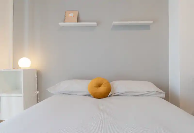Camera da letto luminosa e minimal con letto matrimoniale centrato, pareti grigie, un cuscino rotondo color ocra e una lampada da comodino accesa che crea un'atmosfera calda e moderna.