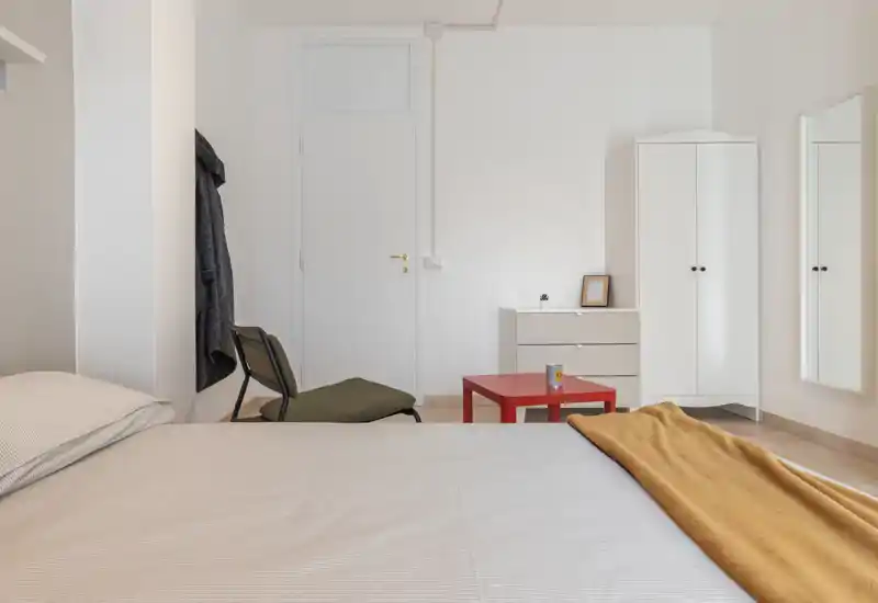 Camera da letto luminosa e minimalista con un ampio letto in primo piano, un armadio e un comò sulla parete di fondo e una coperta calda che aggiunge colore.