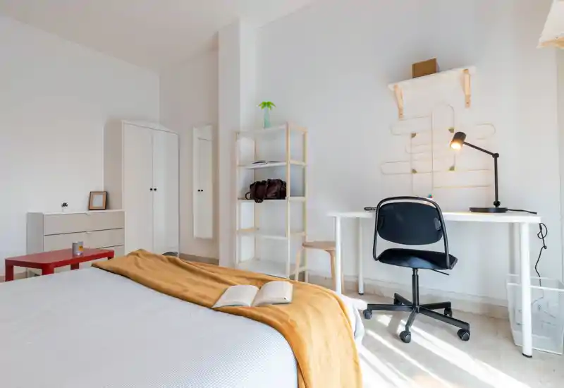 Camera da letto moderna e luminosa con letto ben rifatto, coperta calda e un'area studio funzionale vicino alla finestra — ideale per riposo e smart working.