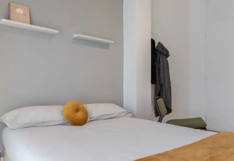 Camera da letto luminosa e minimale con un letto matrimoniale ben rifatto, biancheria morbida e un cuscino decorativo rotondo; toni neutri e mensole semplici creano un’atmosfera rilassante.
