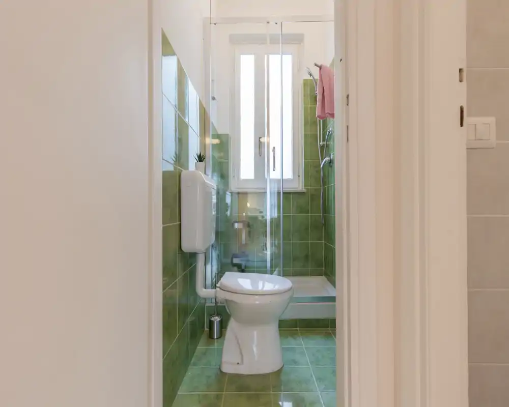 Baño compacto con azulejos verdes en paredes y suelo, inodoro blanco y ducha con mampara frente a una ventana alta. Espacio luminoso y bien cuidado, ideal para un apartamento pequeño.