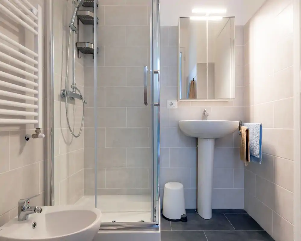 Baño moderno y compacto con mampara de ducha de cristal, lavabo de pedestal, bidé y radiador de pared. Azulejos neutros y líneas limpias ofrecen un aspecto fresco y minimalista, ideal para alquileres de corta estancia.