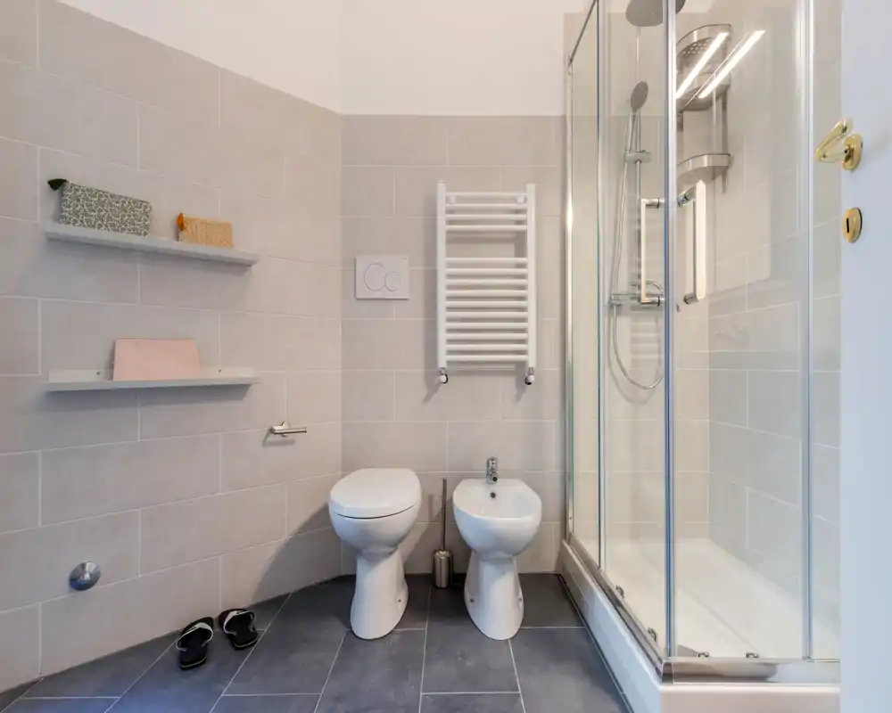 Baño moderno y compacto con inodoro, bidé y ducha con mampara de cristal, azulejos neutros y toallero calefactado.