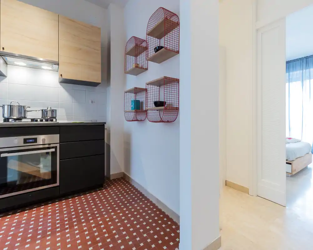 Rincón de cocina moderno y luminoso con horno empotrado, placa de cocina y estantes decorativos en la pared. Suelo de baldosas con patrón y muebles de madera clara crean una sensación acogedora y contemporánea.