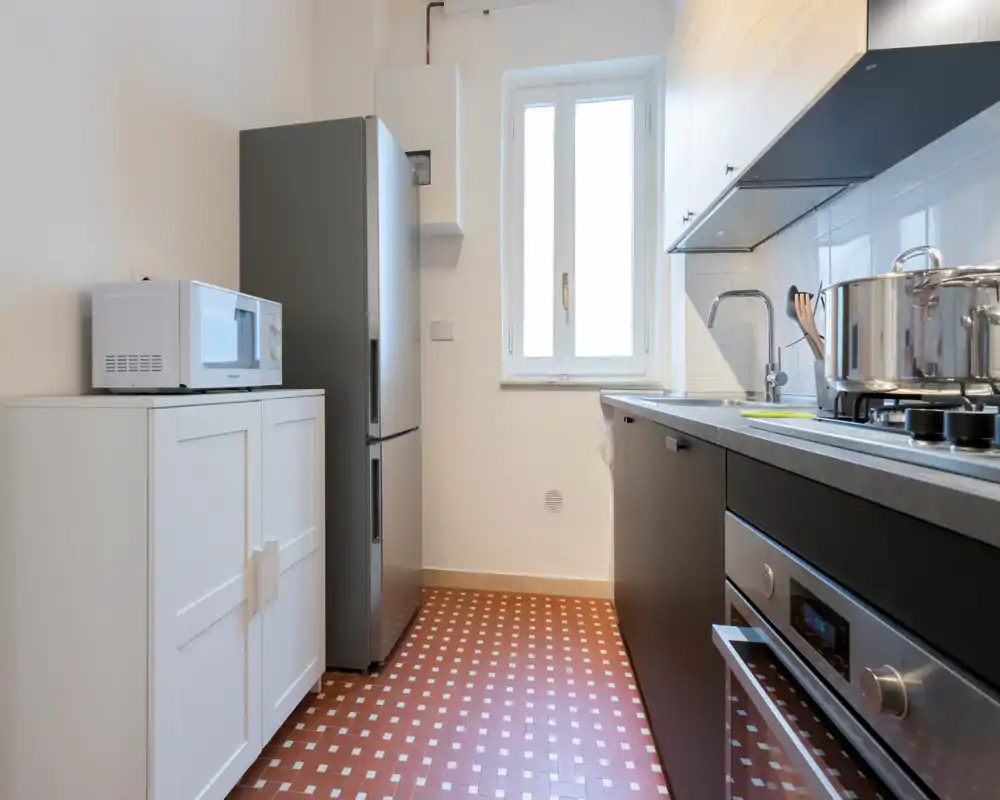 Cocina compacta y moderna con horno empotrado, frigorífico de acero inoxidable, microondas y placa de gas; la luz natural de la ventana realza la encimera y las ollas ordenadas.