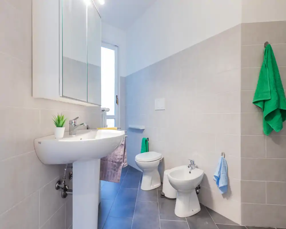 Baño moderno y limpio con lavabo de pedestal, mueble con espejo, inodoro y bidé. Azulejos claros y luz natural desde la ventana crean un ambiente luminoso y fresco.