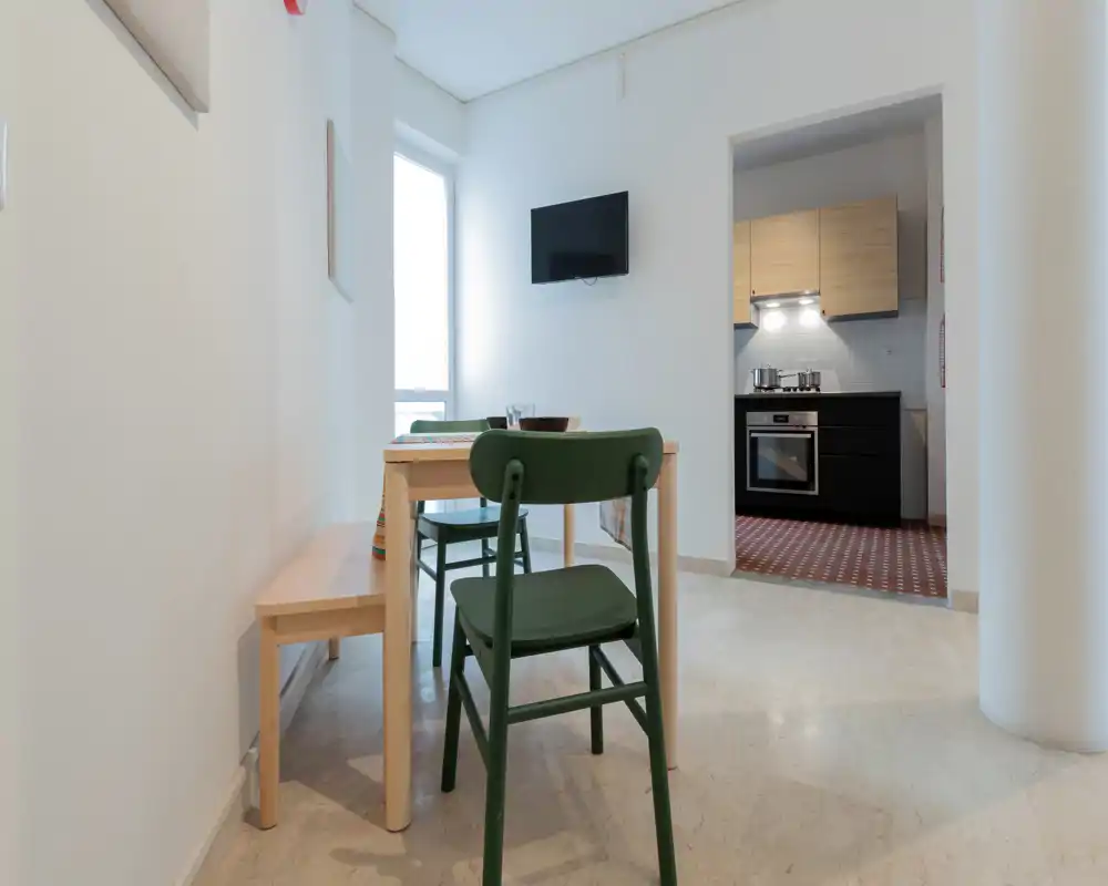 Zona de comedor compacta con una mesa de madera sencilla, un banco y sillas verdes, conectada a una pequeña cocina abierta. Espacio luminoso y minimalista, ideal para comidas informales.