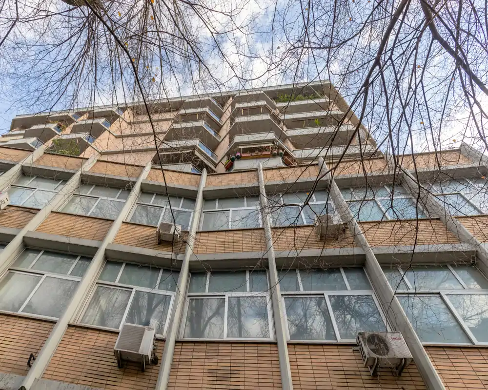 Vista exterior de un edificio residencial de varias plantas tomada desde el nivel del suelo, con balcones, ventanas y unidades de aire acondicionado bajo ramas desnudas.