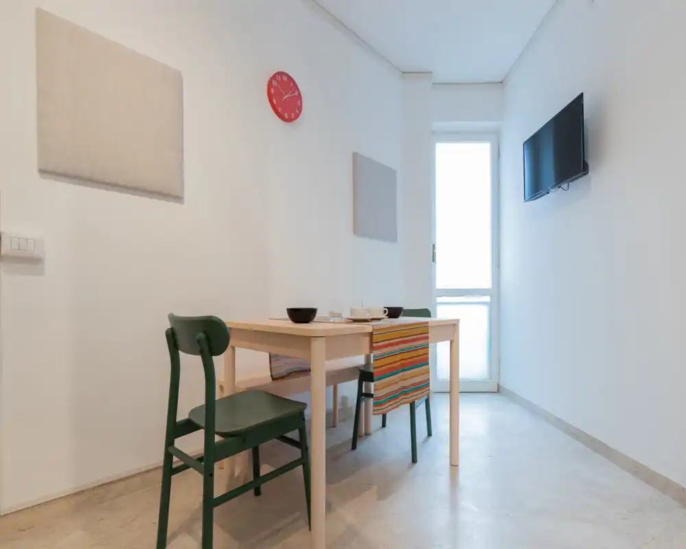 Área de comedor minimalista con una mesa de madera sencilla, dos sillas verdes y un banco, camino de mesa colorido y luz natural desde una puerta de cristal — funcional y luminosa para las comidas.