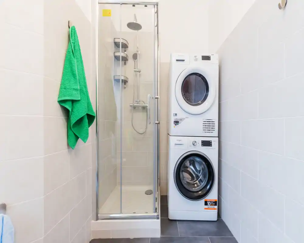 Baño compacto con ducha de mampara de cristal y columna de lavadora-secadora apiladas; azulejos neutros y una toalla verde que aporta un toque fresco.