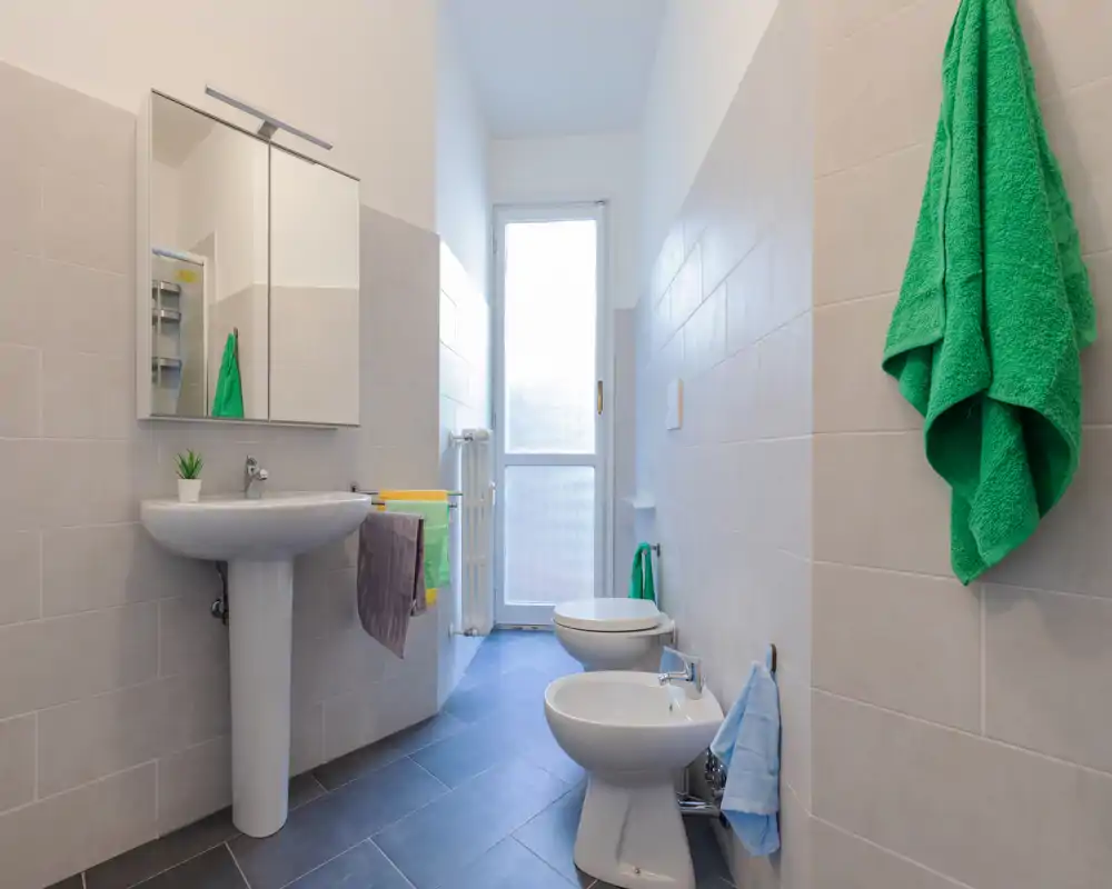 Baño moderno y luminoso con lavabo de pie, bidé y sanitario; azulejos claros y puerta de vidrio esmerilado que aportan sensación de limpieza y amplitud.