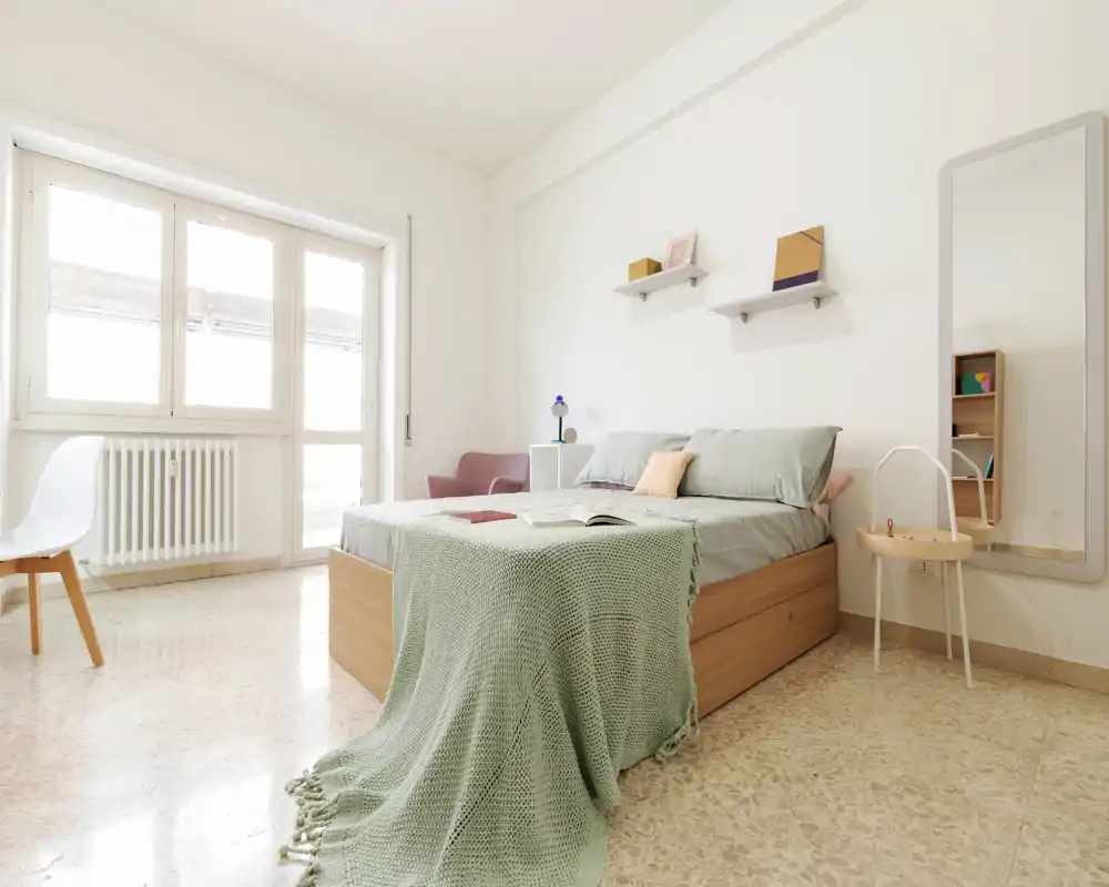 Bright, minimalist bedroom with a full wooden bed, soft green throw and natural light from large windows; airy and inviting for rental listings.