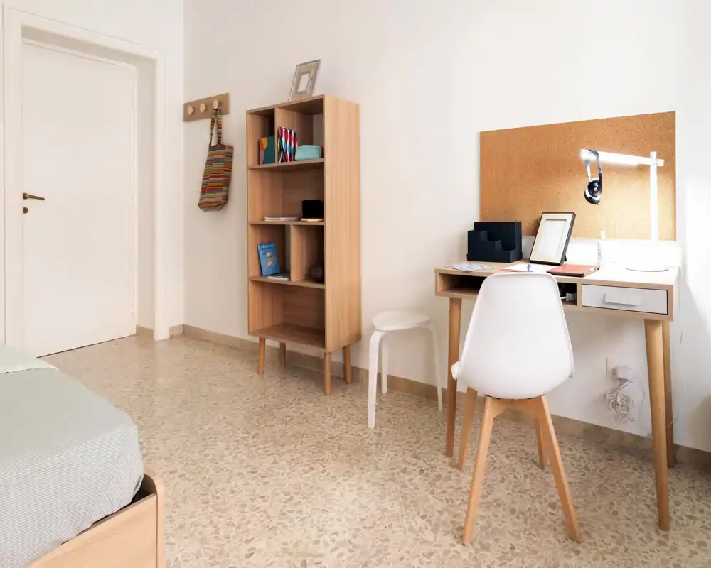 Bright, minimal bedroom with a wooden single bed visible at the left, a compact wooden desk with corkboard and task lamp, and a tidy shelving unit — ideal for a student or single occupant.